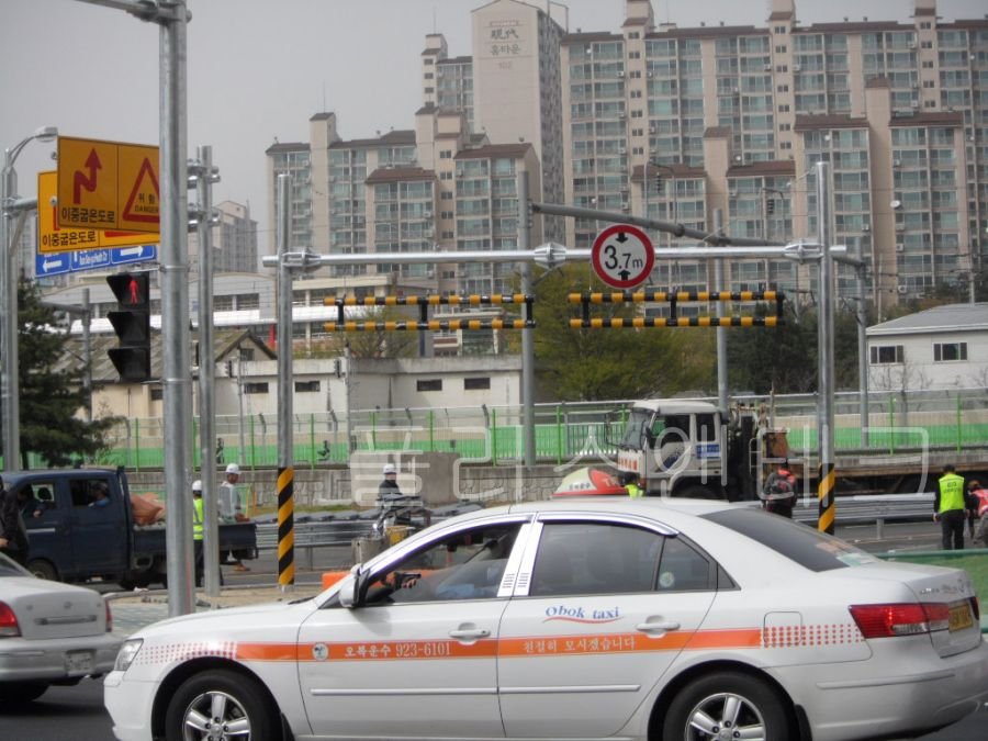 용융아연도금 처리된 은백색 지주에 3.7m 제한 표지판과 충격 흡수용 매달림식 바가 적용된 통과높이제한시설 아래를 택시가 지나가는 모습.
