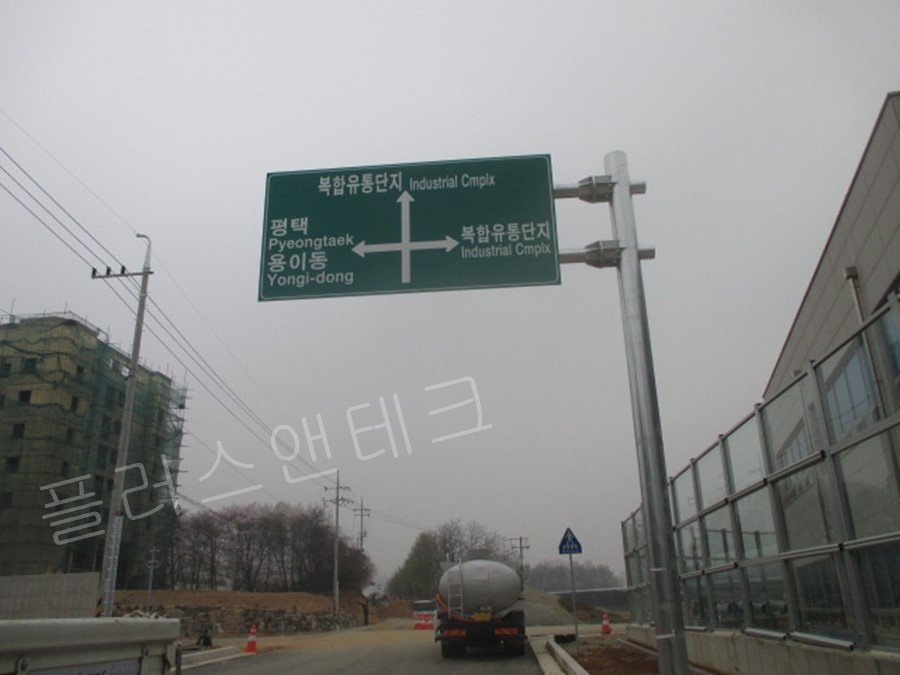 안성 복합유통단지 주 진입로 교차로에 설치된 대형 도로표지판. 직진 및 우회전은 복합유통단지, 좌회전은 평택 및 용이동 방향을 명확하게 안내하며, 대형 탱크로리 차량이 지나가는 현장 모습