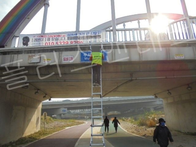  안전 조끼를 착용한 작업자가 무지개다리 하부에서 전동 드릴을 사용하여 콘크리트 천공 및 앙카 설치 작업을 수행하는 장면, 하단에 작업 차량 대기 중
