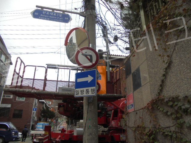 복잡한 통신선과 반사경 사이로 새로운 어린이보호구역 표지판을 조심스럽게 인양하여 위치를 잡고 있는 측면 시공 장면