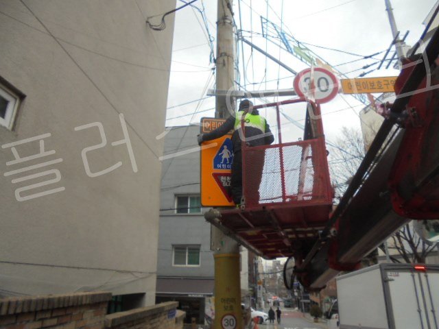 안전 조끼를 입은 전문 작업자가 고소작업차 위에서 밝은 주황색의 스쿨존 30km/h 제한속도 LED 표지판을 조립하고 배선 작업을 하는 디테일한 모습